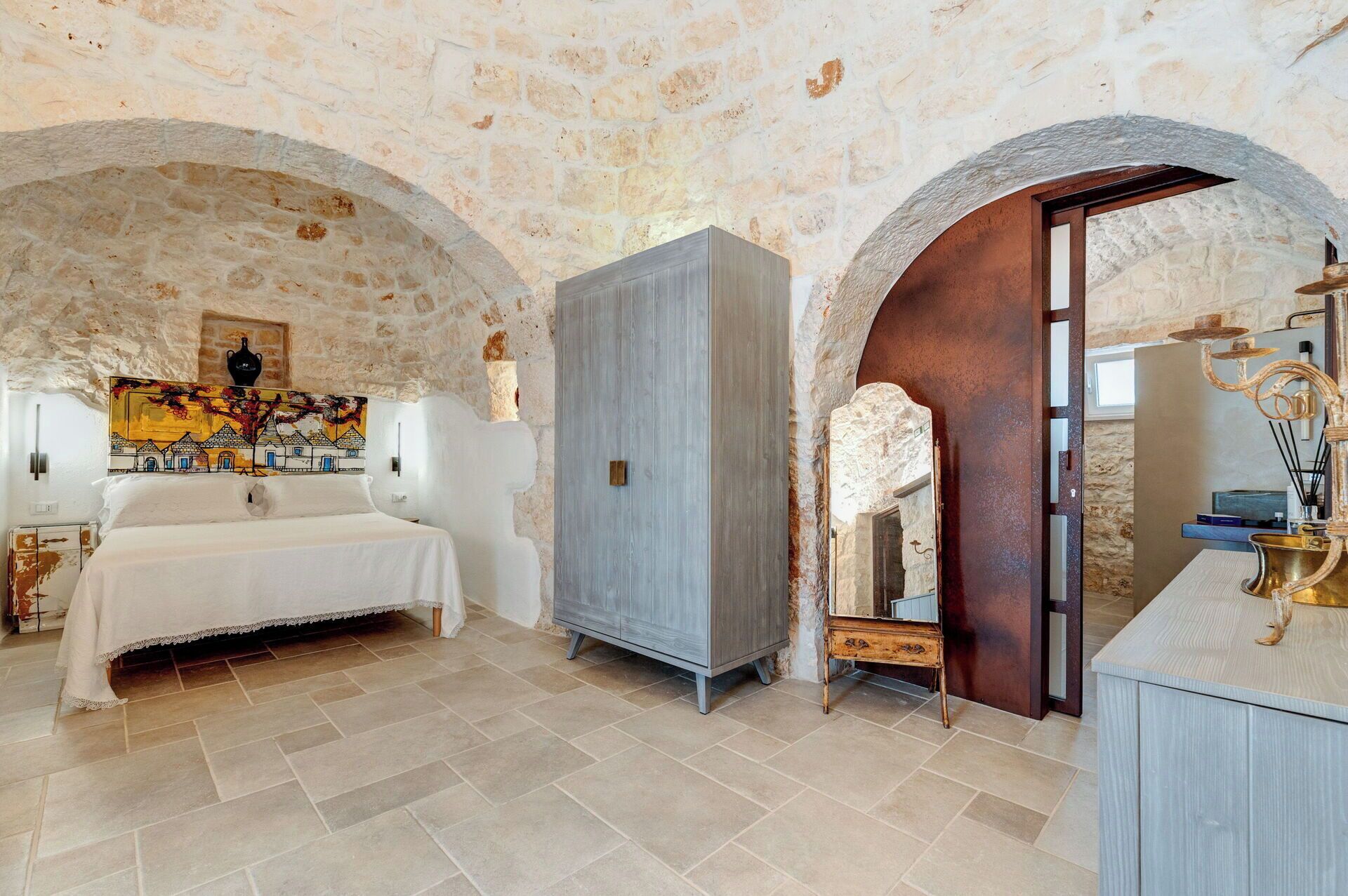 Une pièce comprenant un lit, une armoire et un miroir. Les murs sont en pierre et le sol est carrelé.