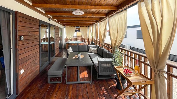 A covered outdoor seating area with a wooden deck, a sofa, a table, and a small table with a bottle and fruit. - Stunning home in Privlaka (Privlaka)