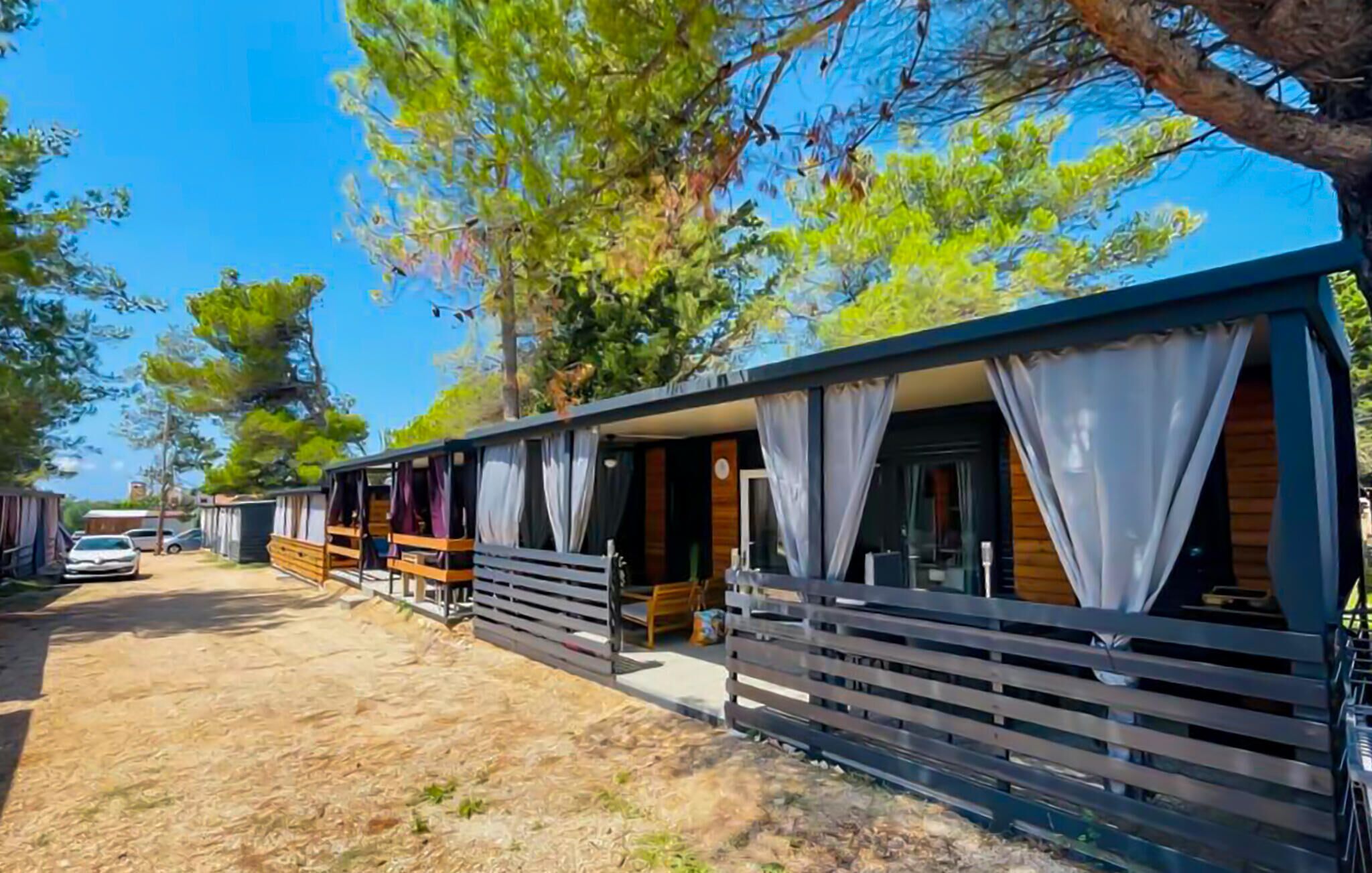 A row of mobile homes with wooden railings and white curtains, a car parked in front, and trees in the background.