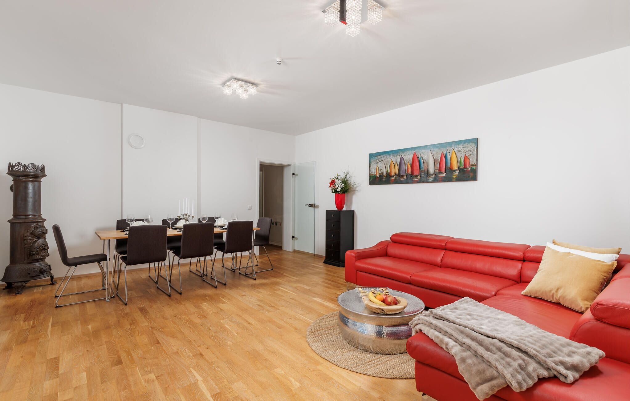 A modern living room with a red sofa, a dining area with a wooden table, and a painting of sailboats on the wall.