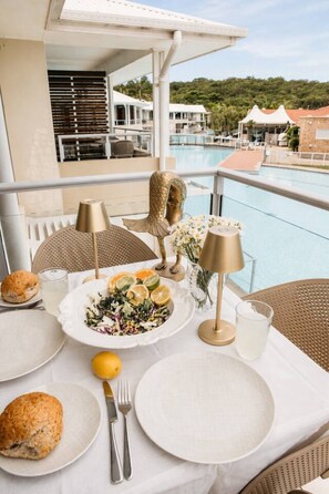 A dining table with plates, cutlery, and a salad, set on a balcony overlooking a pool and buildings. - The Oaks Pacific Blue 1 Bedroom Apartment (Salamander Bay)