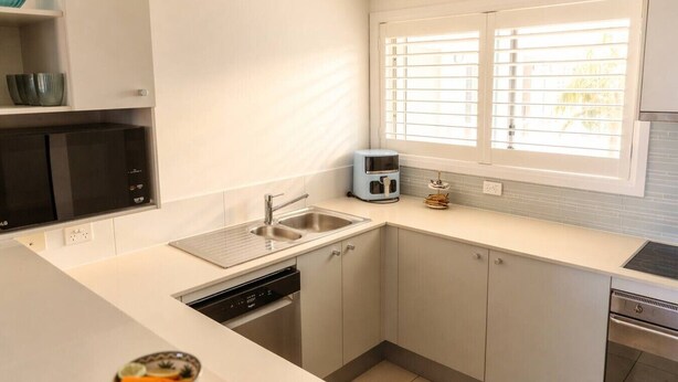 A modern kitchen with a built-in dishwasher, a coffee maker, and a window with blinds. - The Oaks Pacific Blue 1 Bedroom Apartment (Salamander Bay)