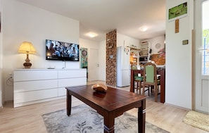 A living room with a TV, a wooden table, a sofa, and a kitchen area in the background.