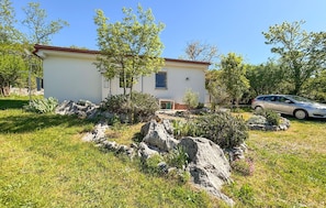 A single-story building with a white exterior and a red roof, surrounded by a well-maintained garden with rocks and shrubs, and a parked car in the background.