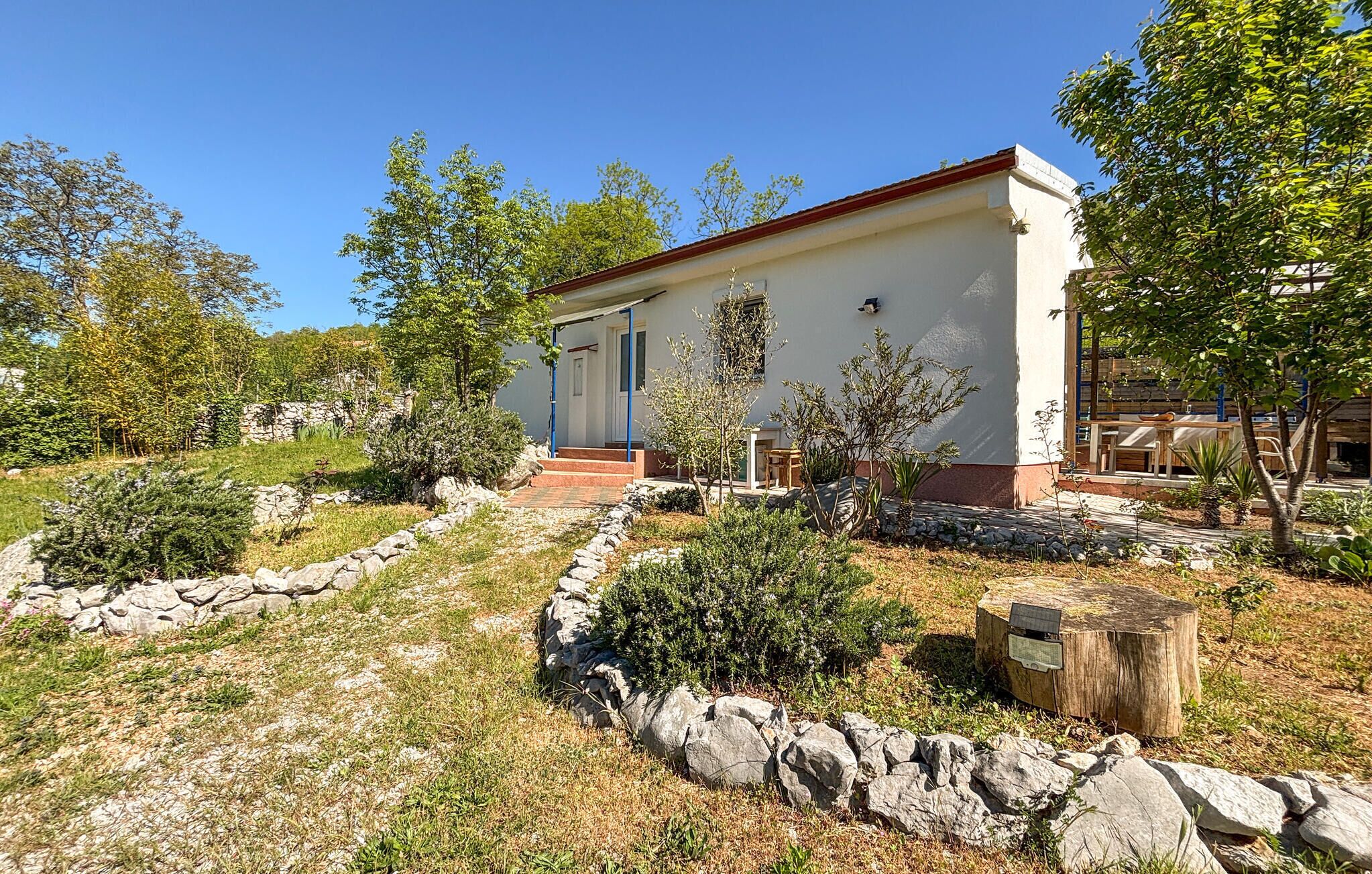 A single-story building with a red roof, surrounded by a garden with various plants and a stone wall.