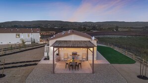 A covered patio area with a table and chairs, a barbecue grill, and a view of a vineyard in the background. - Covered Pavilion + BBQ | Paso Hillside + Vineyards View | Bombay Chardonnay (Paso Robles)