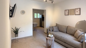 Een woonkamer met een bank, een klein rond tafeltje, een potplant en een keuken zichtbaar door een open doorgang.