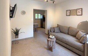 Una sala de estar con un sofá, una mesita redonda, una planta en maceta y una cocina a la vista a través de un vano abierto.