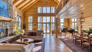 Een interieur van een blokhut met een open haard, een piano, een eethoek en een groot raam met uitzicht op een bergachtig landschap.