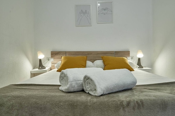 A neatly made bed with two pillows, a blanket, and two rolled towels. There are two bedside lamps and two framed pictures on the wall.