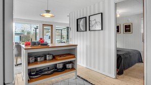 A modern kitchen with white cabinetry, a wooden countertop, and a built-in shelf with cookware. There are two framed pictures on the wall, and a bed with a checkered blanket in the adjacent room.
