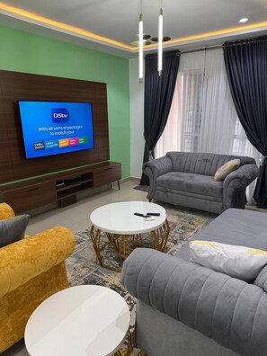 A modern living room with a flat-screen TV mounted on a wooden wall, grey sofas, a yellow armchair, and a white coffee table. - Vine Apartment (Abuja)