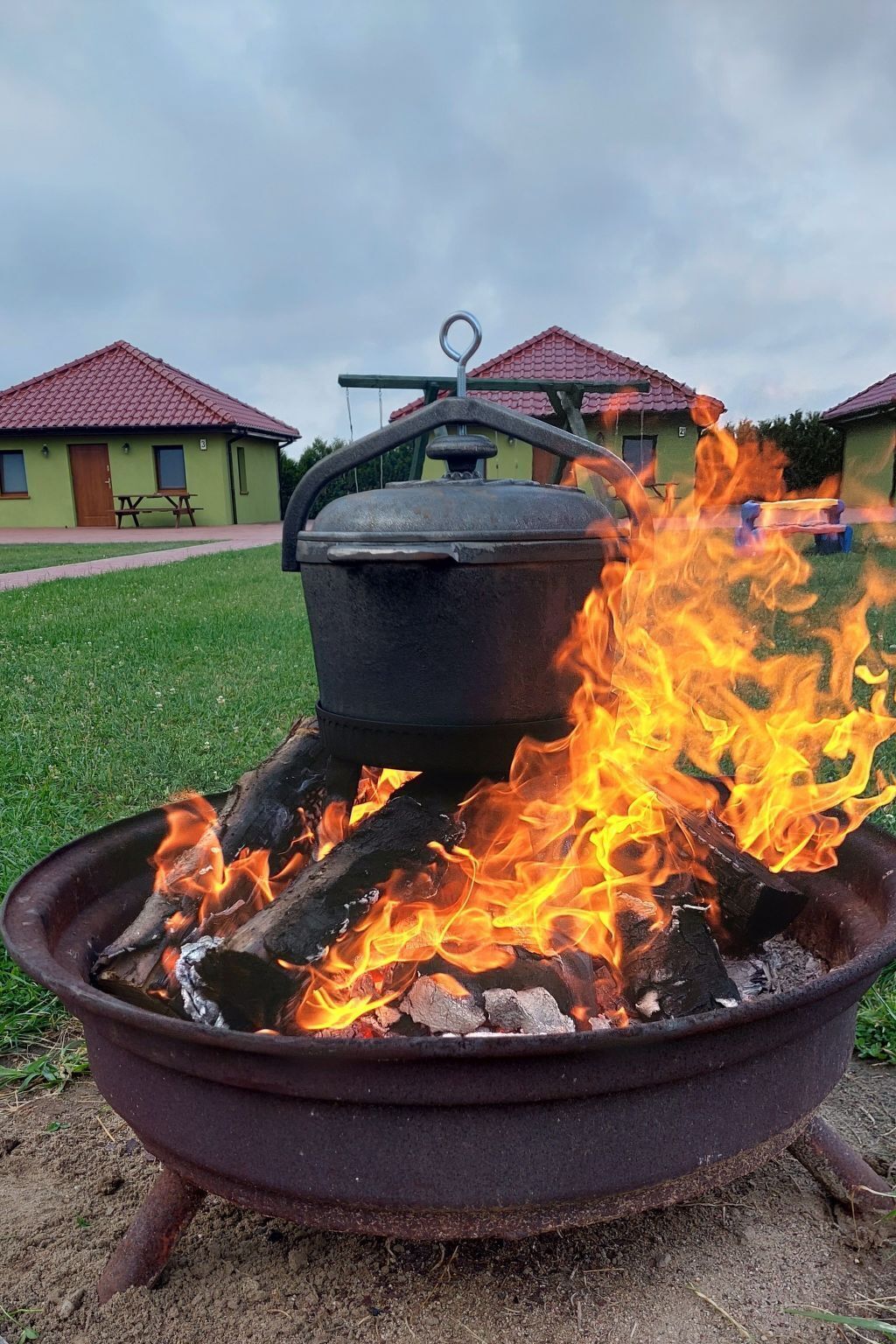 Un ensemble de cuisson portatif pour l’extérieur, avec une marmite posée sur le dessus, entourée de flammes, installé sur une zone herbeuse avec des bâtiments en arrière-plan.