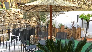 Ein Terrassenbereich mit einer Steinmauer, einem Metallzaun, einem Holzzaun und einem Reetdachschirm.