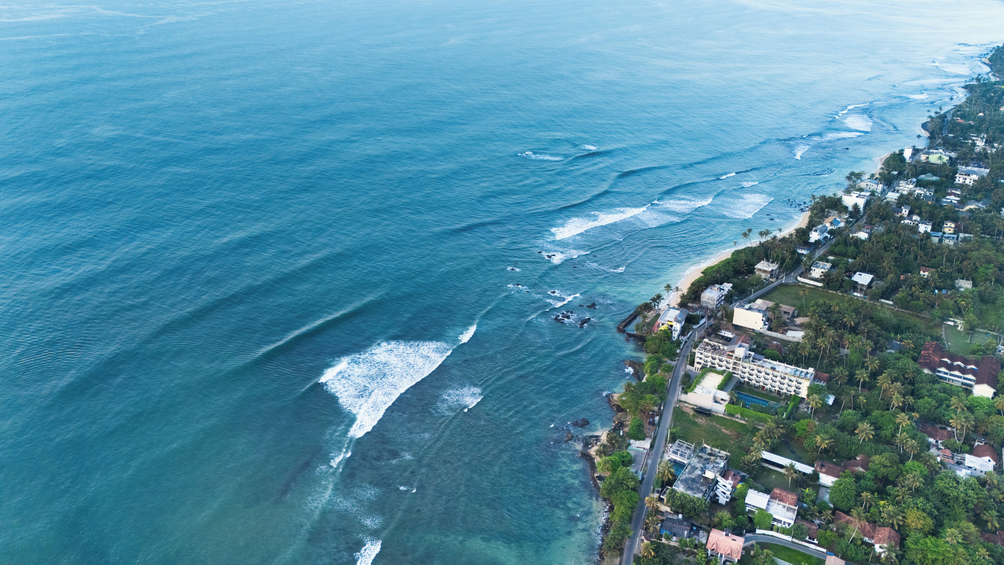 Vista aérea de una zona costera con playa, olas del océano y una área residencial con edificios y vegetación.