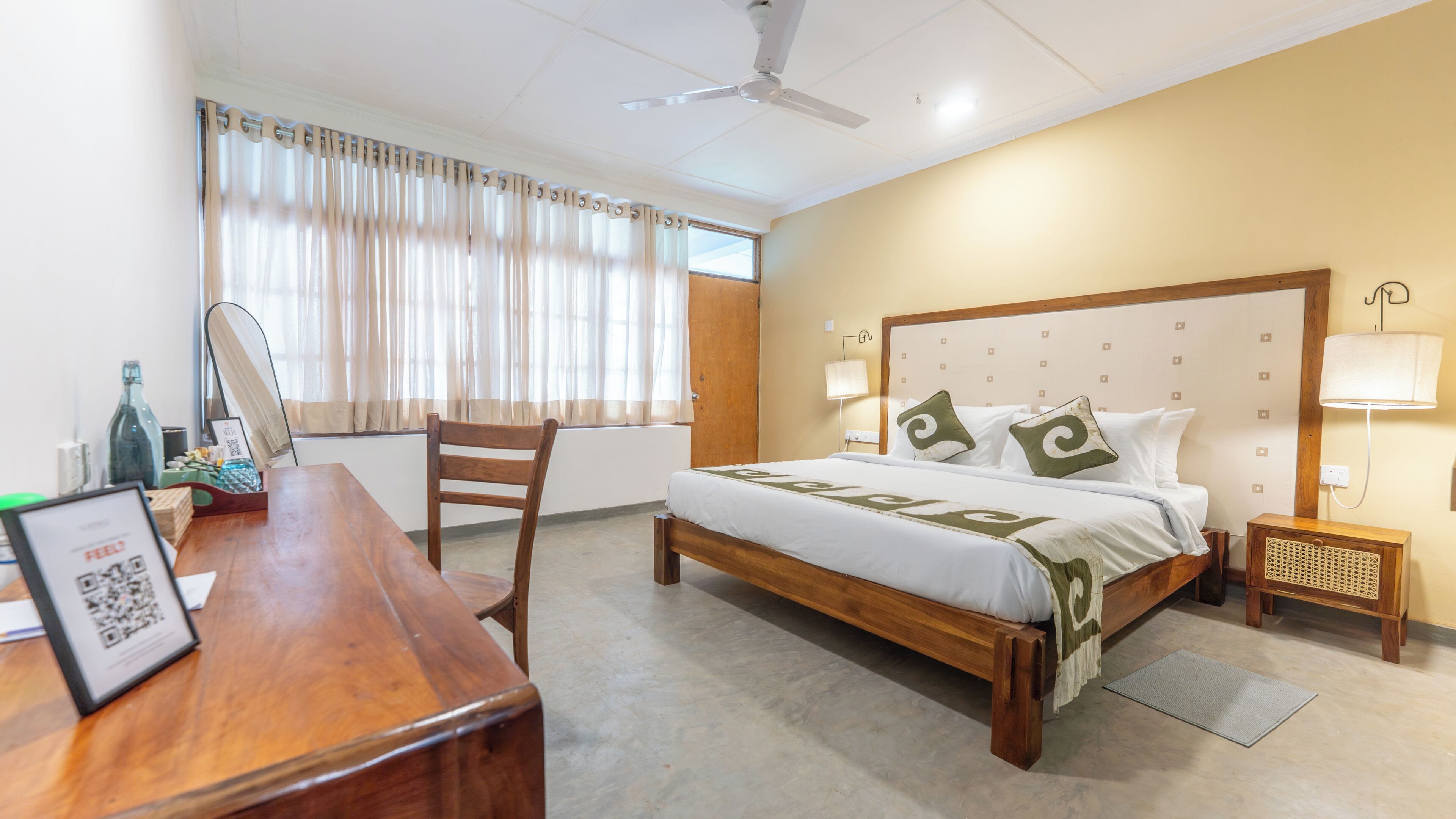 Una habitación de hotel con una cama de madera, un escritorio, una silla, una lámpara de pie y un ventilador de techo.
