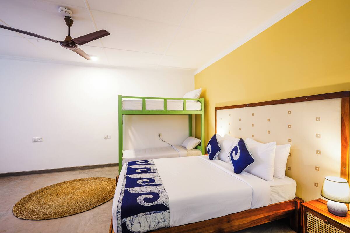 A room with a bunk bed, a single bed, a ceiling fan, a lamp, and a woven mat on the floor.