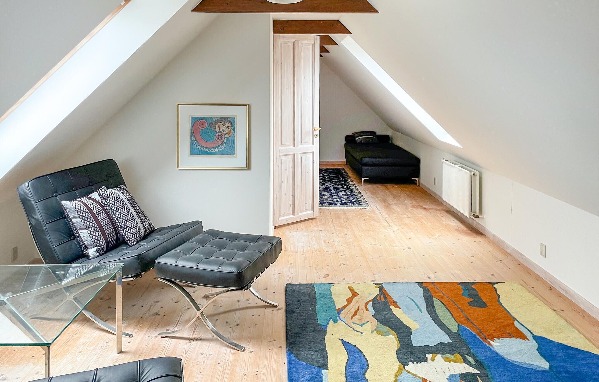 A loft-style room with a high ceiling, a black leather chair, a glass table, a colorful rug, and a black sofa.