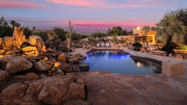 A pool surrounded by rocks and cacti, with a sunset in the background. - 5BDR Oasis w/ Waterfall Pool, Hot Tub, Desert View | Desert Cascase by AvantStay (Carefree)