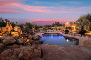 A pool surrounded by rocks and cacti, with a sunset in the background. - 5BDR Oasis w/ Waterfall Pool, Hot Tub, Desert View | Desert Cascase by AvantStay (Carefree)