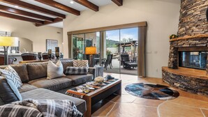 A spacious living room with a stone fireplace, a large sofa, and a dining area with a kitchen in the background. - 5BDR Oasis w/ Waterfall Pool, Hot Tub, Desert View | Desert Cascase by AvantStay (Carefree)