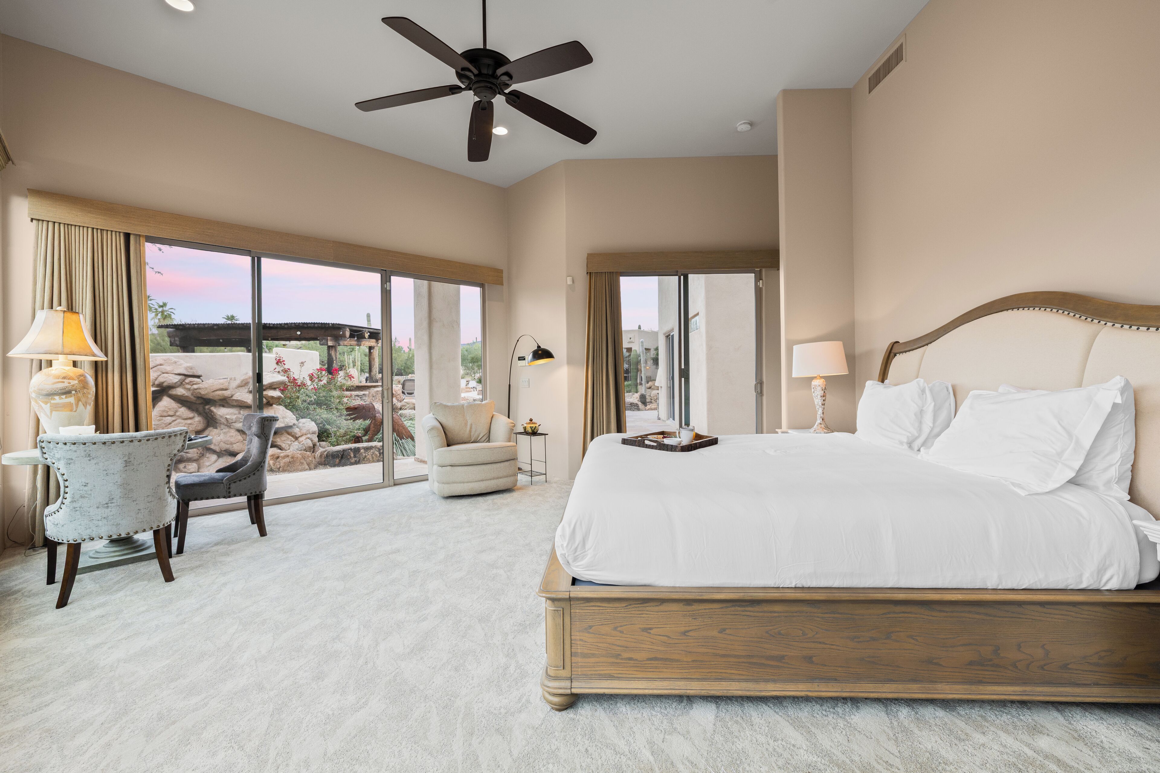 A spacious bedroom with a large bed, a ceiling fan, and a view of a rocky landscape through the large windows.