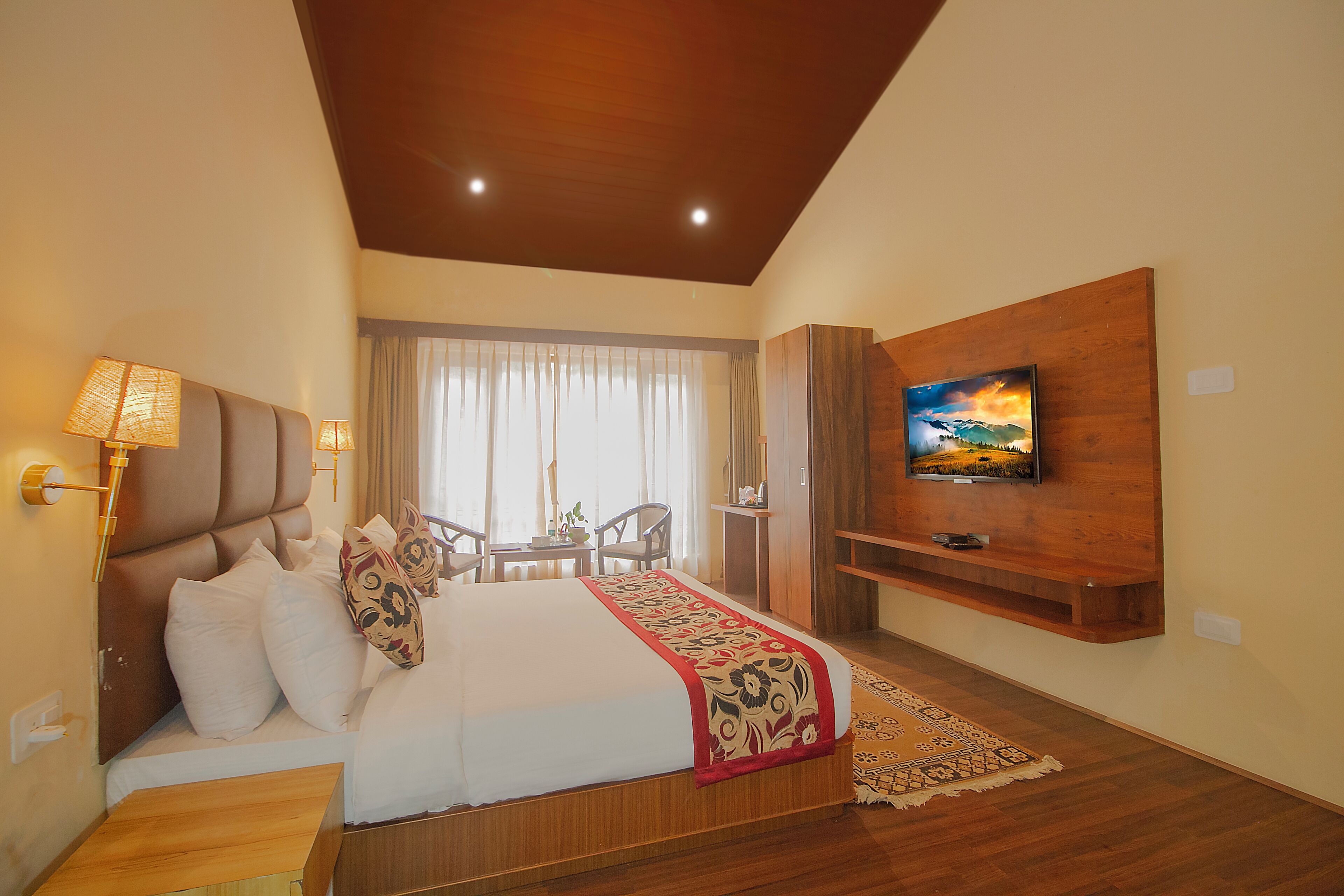 A hotel room with a bed, a TV mounted on the wall, a wooden headboard, and a view of the outdoors through the window.