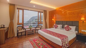 Habitación de hotel con una cama grande, paredes de madera, vistas a un paisaje montañoso, un escritorio con una silla y una mesita con una planta.