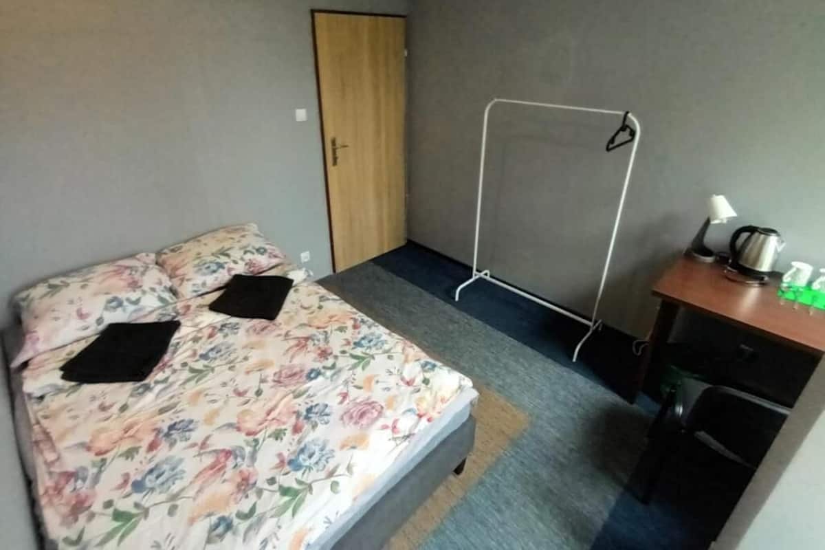 Ein kleines Zimmer mit einem Bett, einem Schreibtisch und einem Stuhl. Eine Tür und ein Fenster sind vorhanden.