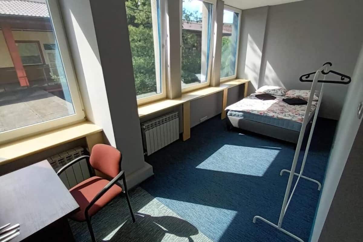 Ein kleines Zimmer mit einem Bett, einem Schreibtisch und einem Stuhl. Große Fenster sorgen für natürliches Licht und einen Ausblick nach draußen.