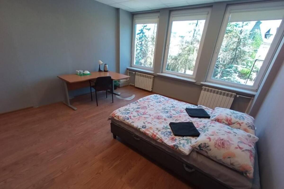 Ein Schlafzimmer mit Bett, Schreibtisch und Stuhl. Drei Fenster sorgen für natürliches Licht.