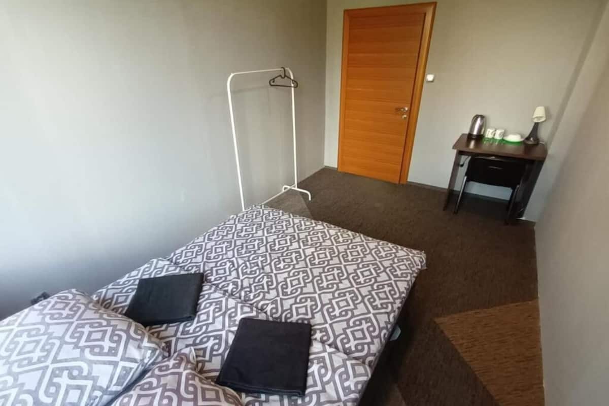 Ein kleines Zimmer mit einem Einzelbett, einer Bettdecke mit Muster, einem Nachttisch mit Lampe und einer Holztür.