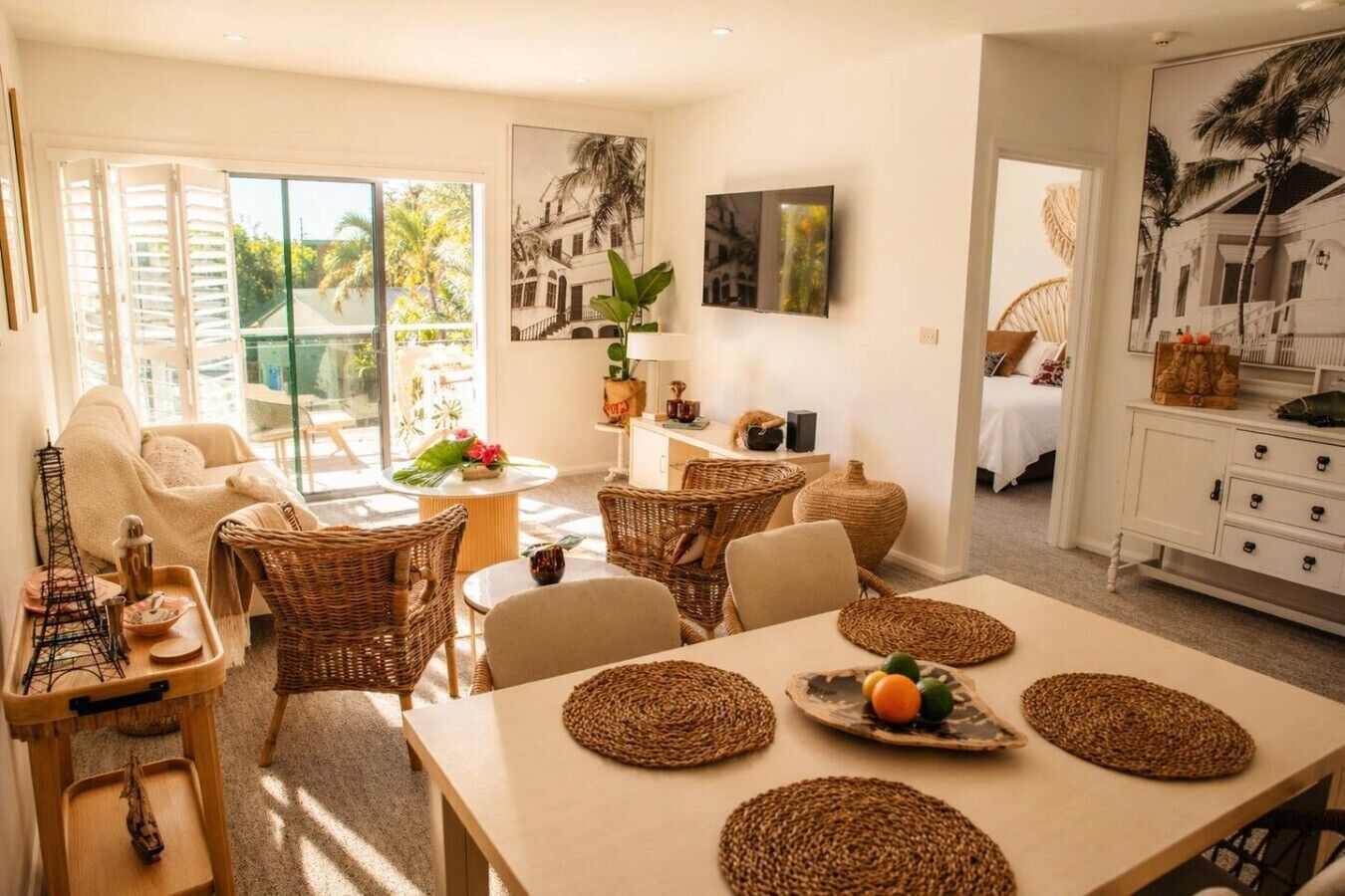 A well-lit room with a dining table, wicker chairs, and a sofa. There is a large window providing natural light and a view of greenery outside.