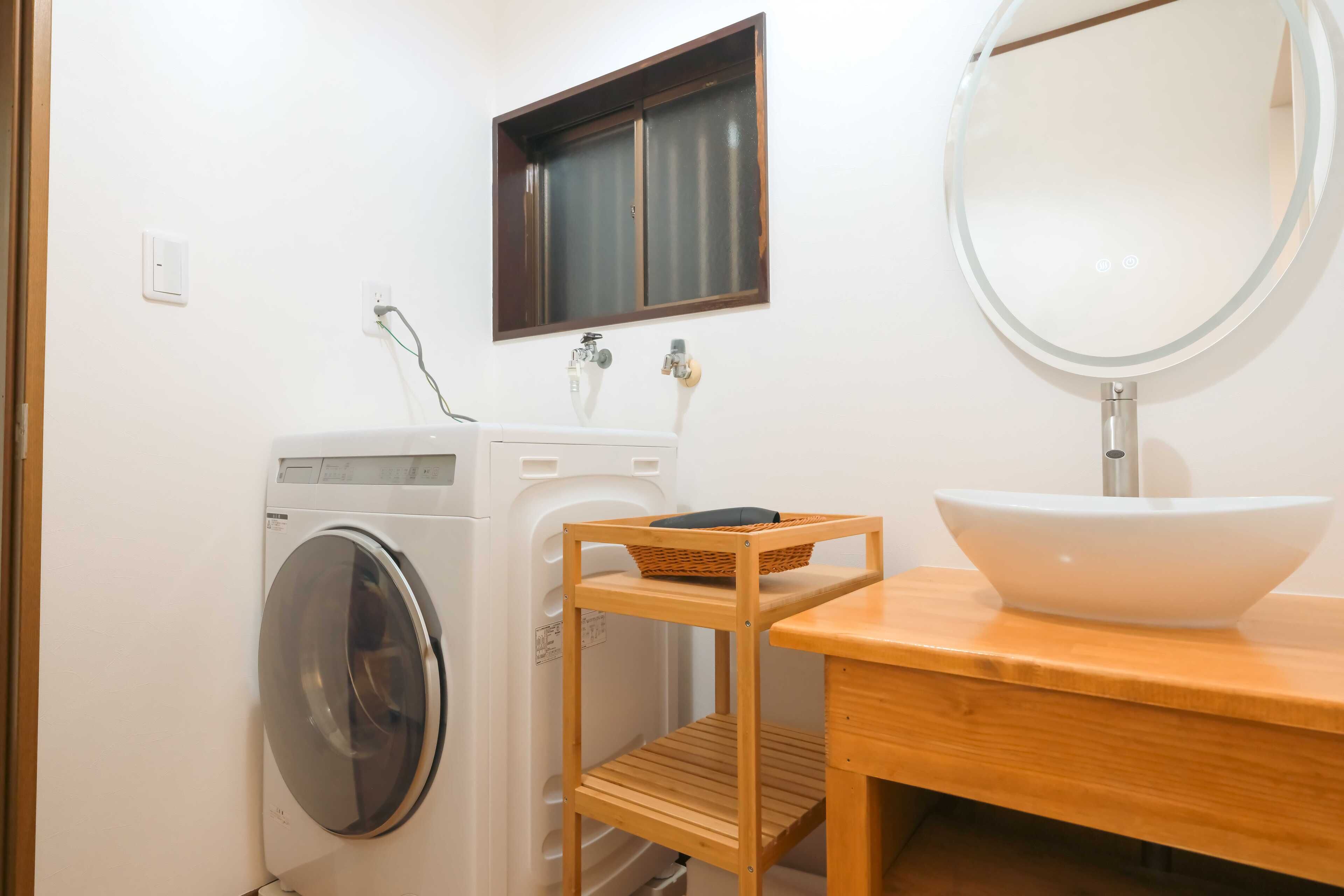 A compact bathroom with a washing machine, a small window, a wooden shelf, and a round mirror.