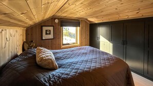 A bedroom with a wooden ceiling, a bed with a quilted bedspread, a pillow, a framed picture on the wall, and a window with a view of trees. - Amazing home in Røn with sauna (Røn)