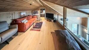 A spacious loft-style room with a high ceiling, wooden beams, a large window, a sofa, a TV, and a rug. - Amazing home in Røn with sauna (Røn)
