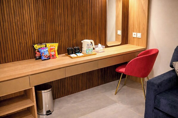 Una habitación de hotel moderna con un panel de pared de madera, un escritorio de madera grande con aperitivos, una cafetera y una silla roja.