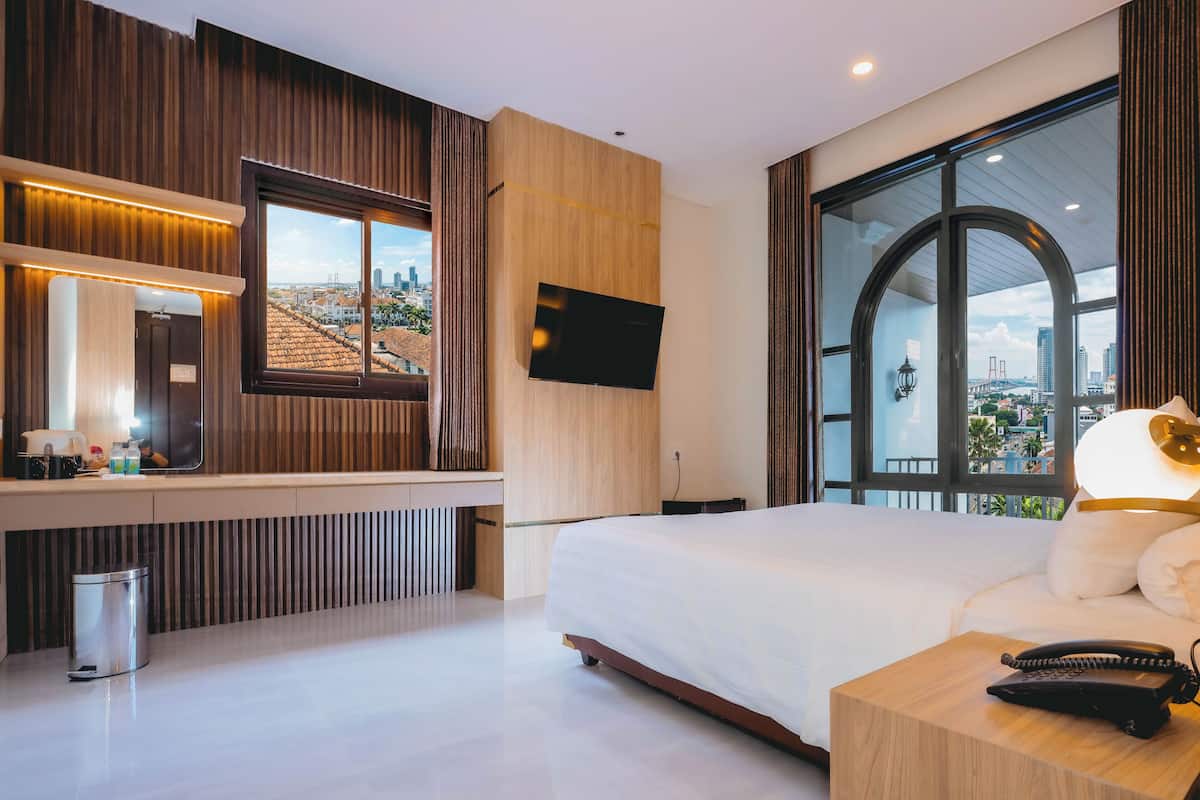 Une chambre d’hôtel moderne dotée d’un grand lit, d’une table de chevet avec un téléphone, d’un téléviseur fixé au mur et d’une grande fenêtre donnant sur la ville.