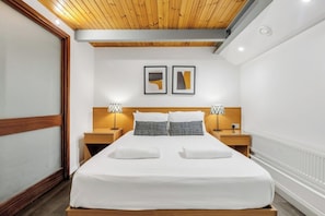 A hotel room with a wooden ceiling, a large bed with white bedding, two bedside tables with lamps, and two framed pictures on the wall.