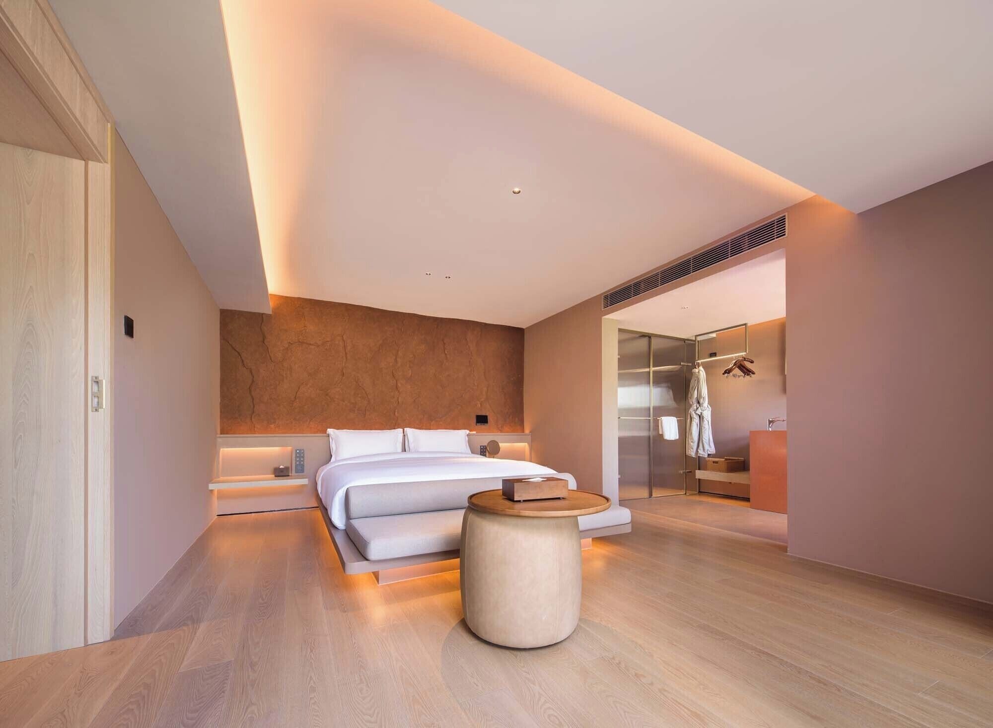 A modern hotel room with a large bed, a round wooden side table, a walk-in closet, and a glass shower area.