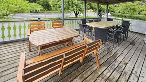 Eine hölzerne Terrasse im Freien mit einem Picknicktisch und Bänken, umgeben von Stühlen und einem überdachten Bereich.