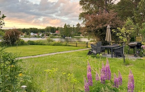 Un espace extérieur verdoyant avec une table et des chaises, un barbecue et un sentier menant à un plan d’eau.