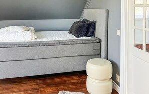 Quarto com sofá-cama cinza, um banquinho redondo branco e piso de madeira.