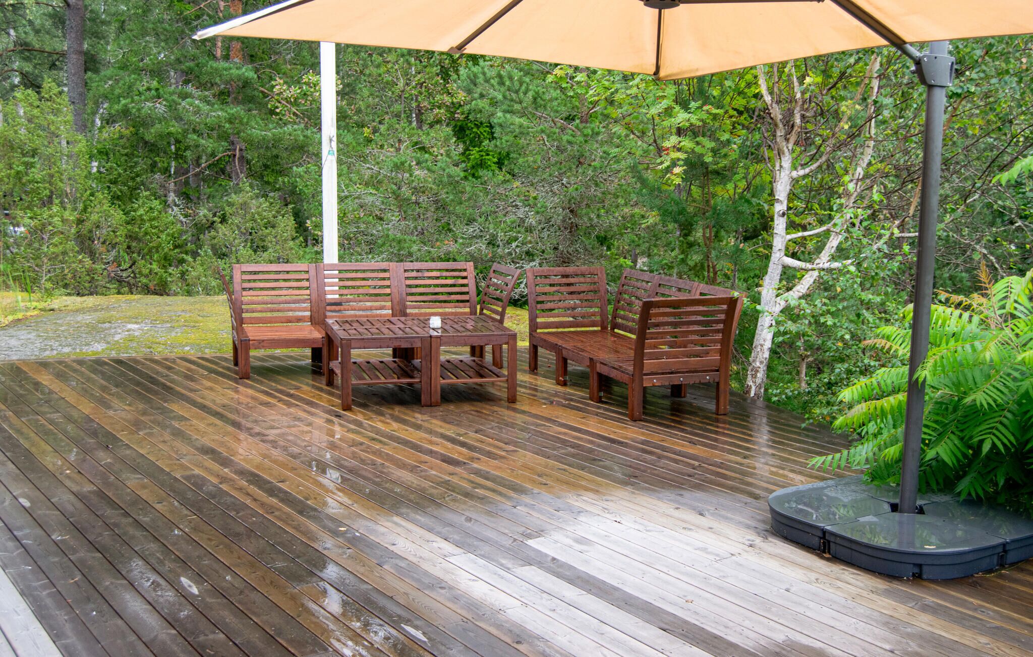 An outdoor wooden deck with a table and chairs under a canopy, surrounded by lush greenery.