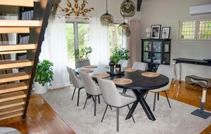 Uma sala de jantar com uma mesa redonda, seis cadeiras e um candelabro. Há plantas em vasos e uma estante de livros ao fundo.