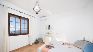 A bedroom with a large window, a bed with a blue pillow, a bedside table with a lamp, and a hanging light fixture. - Villa Baena in Macaca (Torrox)