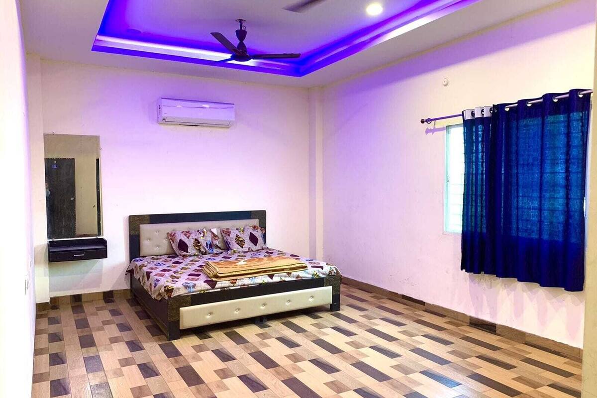 A bedroom with a bed, a ceiling fan, an air conditioning unit, a window with curtains, and a mirror.