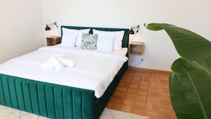 Un lit bien fait, avec une tête de lit verte, du linge de lit blanc et une paire de serviettes. On peut voir deux tables de chevet avec des lampes, et une grande plante verte est également visible.