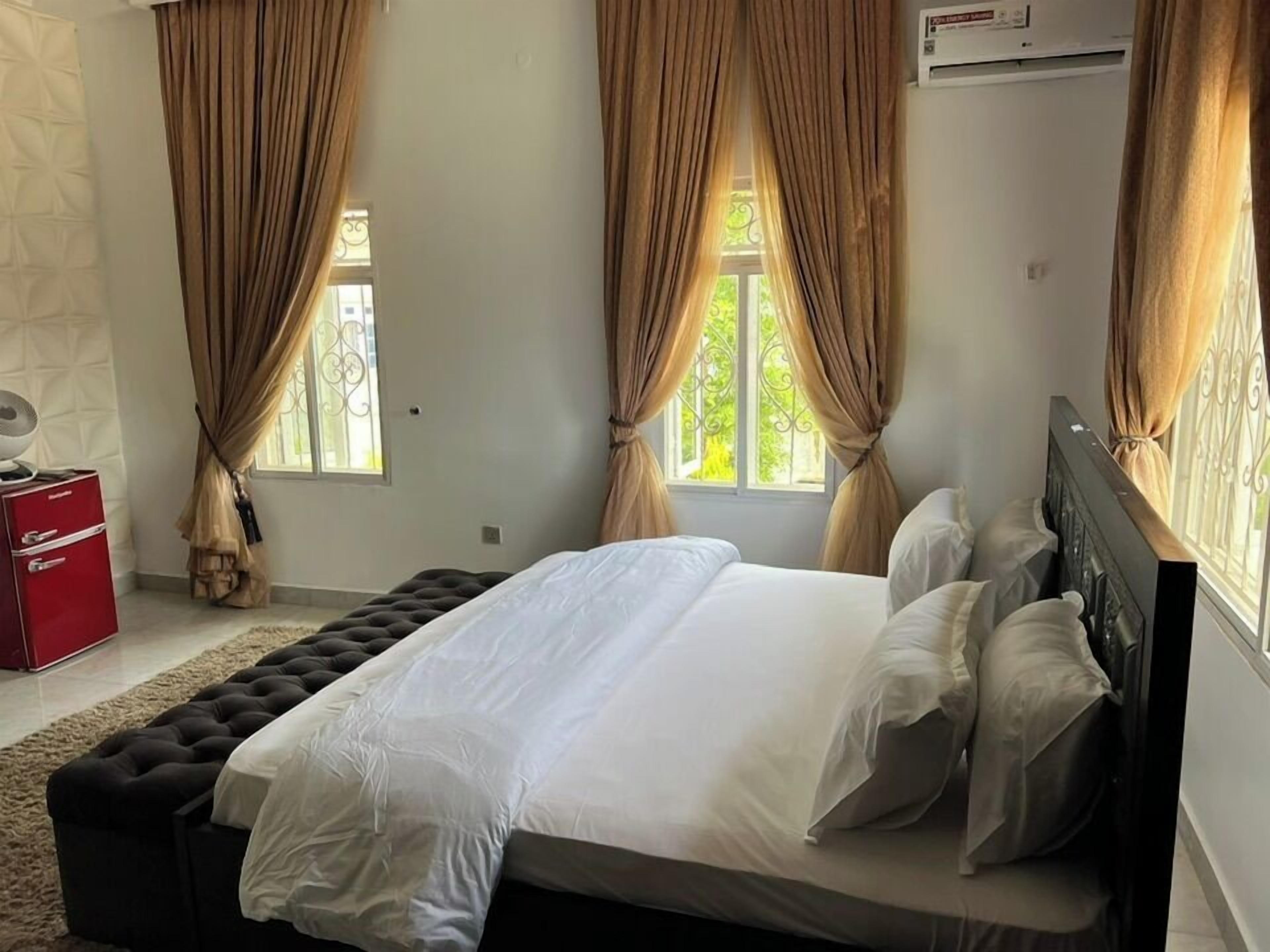 Een slaapkamer met een groot bed, beige gordijnen, een rode kast en een raam met uitzicht op groen.
