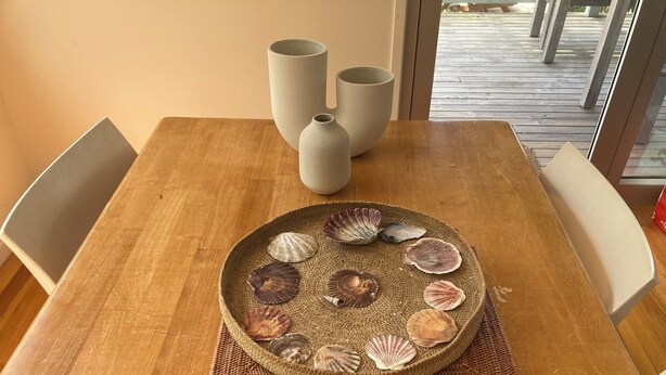 A wooden dining table with a woven placemat holding a decorative plate of seashells. - 20 Bradley (Coles Bay)